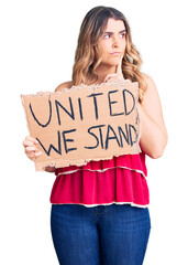 Young caucasian woman holding united we stand banner serious face thinking about question with hand on chin, thoughtful about confusing idea