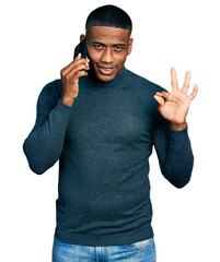 Fototapeta premium Young black man having conversation talking on the smartphone doing ok sign with fingers, smiling friendly gesturing excellent symbol