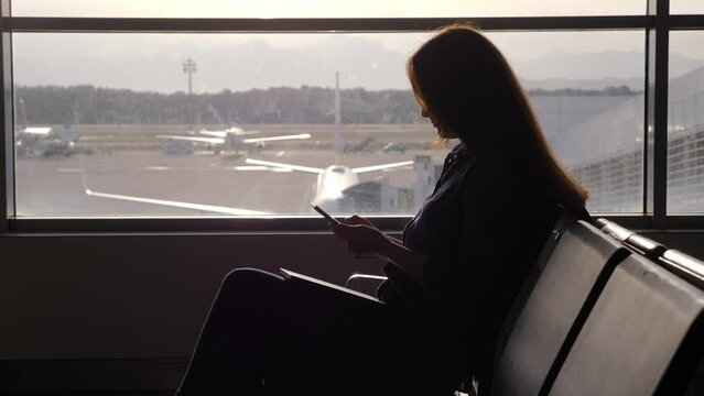 Pensive woman spend time online, using smartphone while wait for boarding. Silhouetted shot of passenger sitting at terminal lounge against window. Blurred apron area and commercial airliners on back