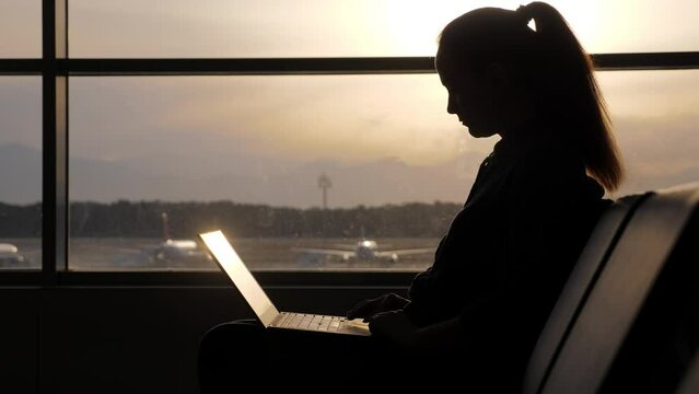Woman continue browsing using laptop, silhouetted shot at airport lounge. Bright evening sun shine through window, blurred background, airliners parked at apron area