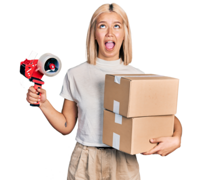 Beautiful young blonde woman holding packing tape and cardboard boxes angry and mad screaming frustrated and furious, shouting with anger looking up.