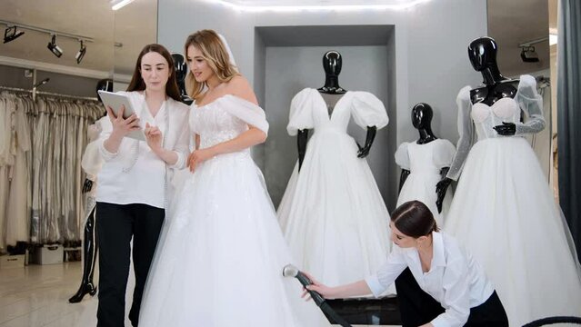 Wedding dress shop owners with digital tablet helping choose a bridal gown and try on a wedding dress at the wedding studio