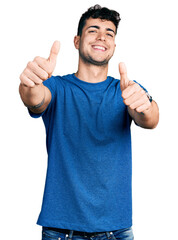 Young hispanic man wearing casual t shirt approving doing positive gesture with hand, thumbs up smiling and happy for success. winner gesture.