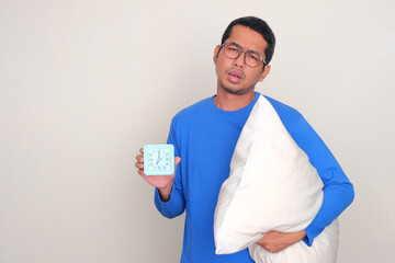 A man with lazy expression holding pillow and showing alarm clock