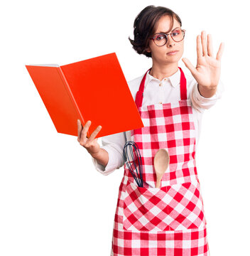 Beautiful young woman with short hair wearing professional baker apron reading cooking recipe book with open hand doing stop sign with serious and confident expression, defense gesture