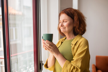 Peaceful senior woman inhaling tea aroma while standing near window at home