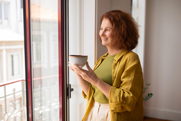 Senior woman drinking cup of coffee while standing near window at home