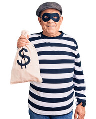 Senior handsome man wearing burglar mask holding money bag looking positive and happy standing and smiling with a confident smile showing teeth