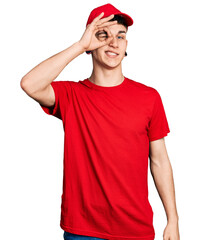 Young caucasian boy with ears dilation wearing delivery uniform and cap smiling happy doing ok sign with hand on eye looking through fingers