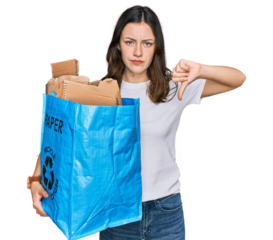 Young beautiful woman holding recycling wastebasket with paper and cardboard with angry face, negative sign showing dislike with thumbs down, rejection concept