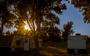 Rv trailer parked at campsite under trees sunburst and open field and forrest 