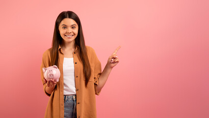Smiling teen girl holding piggy bank and pointing at copy space
