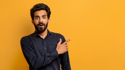 Shocked indian guy pointing aside at copy space on yellow studio background