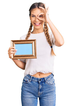 Beautiful caucasian woman with blonde hair holding empty frame smiling happy doing ok sign with hand on eye looking through fingers