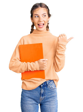 Beautiful caucasian woman with blonde hair holding book pointing thumb up to the side smiling happy with open mouth
