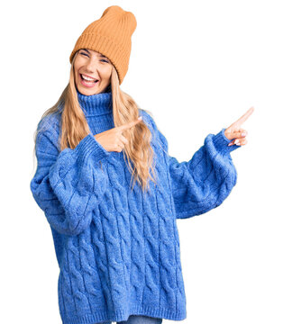 Beautiful caucasian woman with blonde hair wearing wool sweater and winter hat smiling and looking at the camera pointing with two hands and fingers to the side.