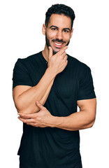 Hispanic man with beard wearing casual black t shirt looking confident at the camera smiling with crossed arms and hand raised on chin. thinking positive.