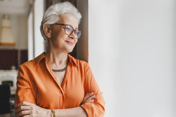 Side view of pretty charming grandmother in orange shirt and glasses leaning against wall with...