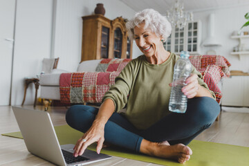 Healthy slim senior female in leggings training at home on mat in front of laptop, watching online tutorial workout, smiling at screen sitting with crossed legs and plastic bottle
