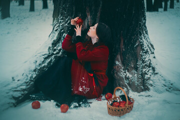 A beautiful young woman in a folklore costume and pigtails is sitting near an old oak tree with a basket of red apples on a snowy winter day. Slavic fairy tale and traditions.