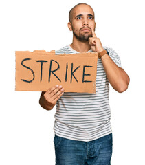 Hispanic adult man holding strike banner cardboard serious face thinking about question with hand...