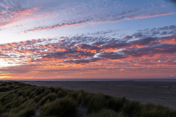 Panoramic sunset view over the dunes on the island of Texel in the Netherlands with long bands of orange coloring clouds in sky with dune grass in forefront and people walking on the wide beach