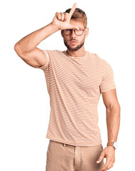 Young caucasian man wearing casual clothes and glasses making fun of people with fingers on forehead doing loser gesture mocking and insulting.