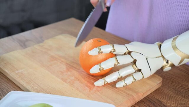 A woman with a prosthetic arm cuts an orange fruit using a kitchen knife in the kitchen. A woman with disabilities tries to cut an orange with one hand, using a prosthetic to help