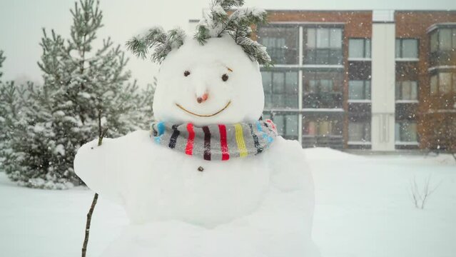 Snowman in a warm scarf during heavy snowfall against the backdrop of a modern residential building and pine trees. The weather outside is frightful, but a cool snowman makes life more fun
