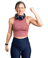 Beautiful caucasian young woman wearing gym clothes and using headphones angry and mad raising fist frustrated and furious while shouting with anger. rage and aggressive concept.