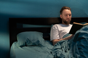 Handsome caucasian man relaxing in bed reading bestseller novel book. Hobbies and free time