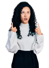 Young hispanic woman with curly hair wearing casual sweater amazed and surprised looking up and pointing with fingers and raised arms.