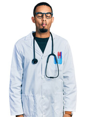 Young african american man wearing doctor uniform and stethoscope making fish face with lips, crazy...