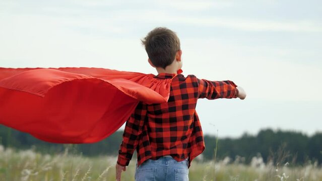 Boy kid plays superhero in red cape, childhood dream. Happy child playing superhero against sky. Little hero in red cloak watches sunset. Brave child winner in red raincoat plays in nature. Kid dreams
