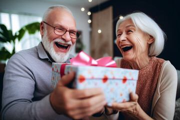 Cheerful senior couple holding gift box and looking at camera while sitting on sofa at home. Selective Focus
