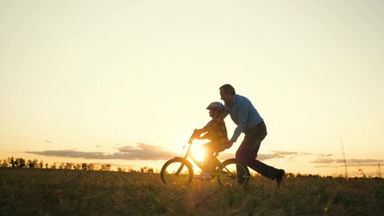 Attentive father silhouette teaches son to ride bike in field at sunset dusk