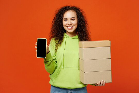 Young woman of African American ethnicity wear green hoody casual clothes hold stack cardboard blank boxes use blank screen mobile cell phone isolated on plain red orange background Lifestyle concept