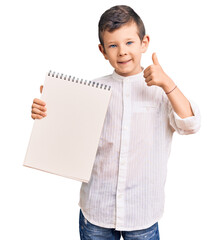 Cute blond kid holding notebook smiling happy and positive, thumb up doing excellent and approval sign