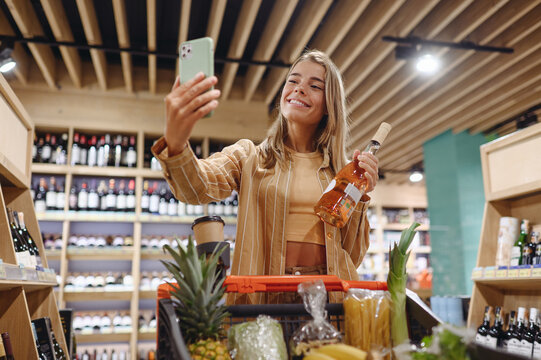 Young customer woman wears casual clothes hold wine bottle do selfie on mobile cell phone shopping at supermaket store shop with trolley cart buy product in hypermarket. Purchasing gastronomy concept.