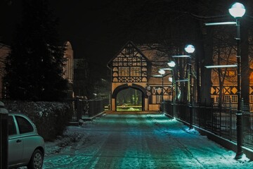 Ulica Pałacowa w miejscowości Iłowa w Polsce. Jest zimowa noc, jezdnię i chodnik pokrywa warstwa śniegu, ciemności rozświetlają latarnie. Ulica zakończona jest bramą w piętrowym budynku.
