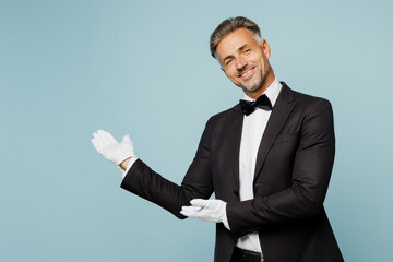 Adult smiling happy barista male waiter butler man wear shirt black suit bow tie elegant uniform point hand aside on area work at cafe isolated on plain blue background. Restaurant employee concept.