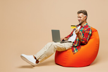 Full body adult man wear red shirt white t-shirt casual clothes sit in bag chair using laptop pc computer hold credit bank card doing online shopping isolated on plain pastel light beige background