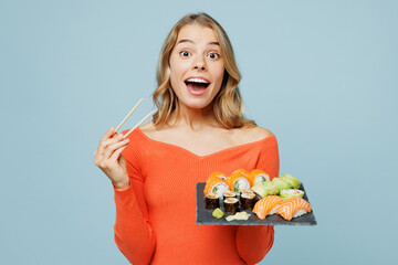 Young surprised happy woman wear orange casual clothes look camera hold eat raw fresh sushi roll served on black plate Japanese food with chopsticks isolated on plain blue background studio portrait.