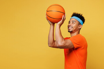 Young fitness trainer instructor sporty man sportsman wear orange t-shirt hold in hand basketball ball shooting training in home gym isolated on plain yellow background. Workout sport fit abs concept.