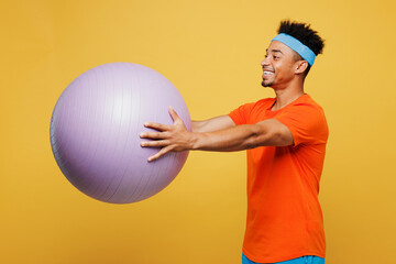 Side view smiling young fitness trainer instructor sporty man sportsman wear orange t-shirt hold in hand fit ball spend time in home gym isolated on plain yellow background. Workout sport abs concept.