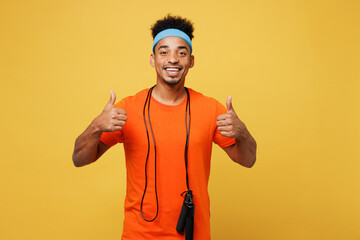 Young fitness trainer instructor sporty man sportsman wears orange t-shirt hold skipping rope show thumb up spend time in home gym isolated on plain yellow background. Workout sport fit abs concept.