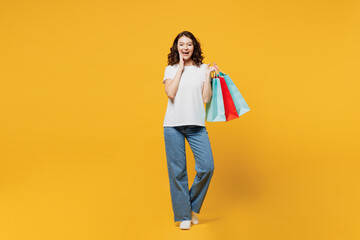 Full body young woman wears t-shirt casual clothes hold in hand paper package bags after shopping look camera hold face isolated on plain yellow orange background. Black Friday sale buy day concept.