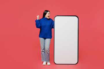 Full body young woman of Asian ethnicity she wear blue sweater casual clothes big huge blank screen mobile cell phone smartphone with area do winner gesture isolated on plain pastel pink background.