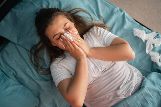Cold and flue concept. Sick woman suffering from running stuffy nose. Upset ill girl lying in bed, blowing her nose using paper napkin. Copy space