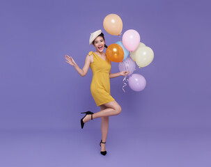 Fototapeta na wymiar smiling happy fun young asian woman in yellow dress celebrating holding bunch of balloons raise up leg isolated on purple background. Birthday and new year party concept